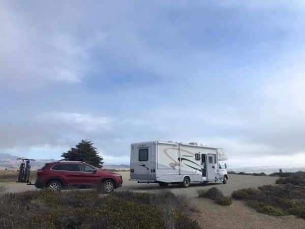 RV is parked in a docking bay at Jalama beach caamping, directly facing the ocean view. A red jeep is parked behind it with bicycles on the tailgate.