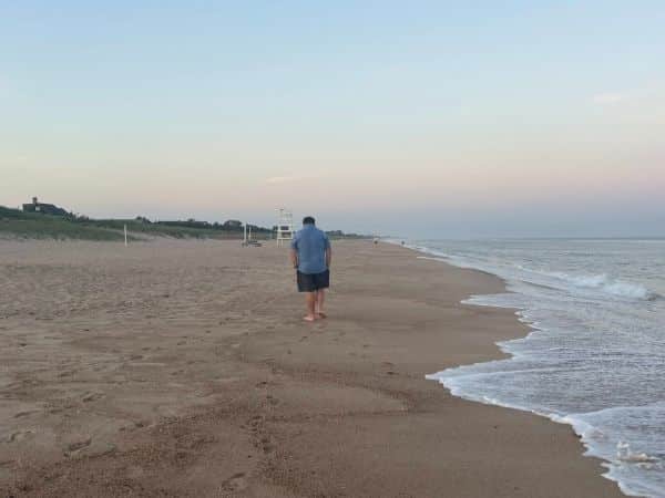 josh has his back to us, walking along atlantic ave beach with a wide sand area and gently lapping waves set aagainst a pale blue sky with hints of pink through the horizon