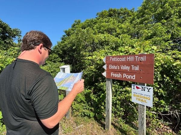 wooden sign post displaying the hiking trails at hither hills state park. Josh stands in front of it checking the map as we get ready to explore the hamptons 