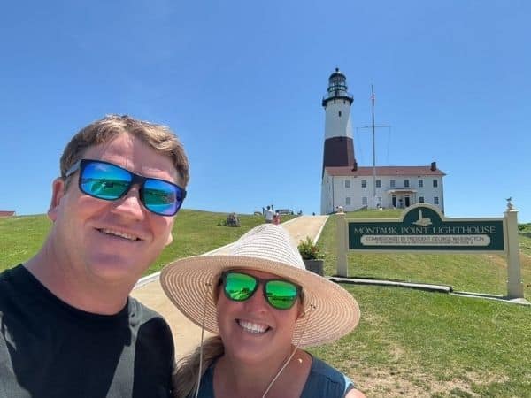 Liz and Josh stand together in the front left corner of the photo. Josh is smiling and Liz wears a straw sunhat. The sky is clear blue and the large white montauk lighthouse stands behind them. 