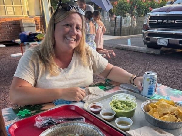 liz sits at an outdoor table at la fondita. She wears a white tshirt and glasses atop her head as she smiles at the camera. The table is covered in food trays hosting tacos and sides, and she holds a can of juice. There are people and a truck in the background. 