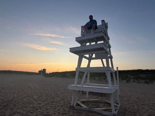 sunset at the hamptons beach. a large whie lifeguard chair stands on the sand, josh sits at the top, orange tinges the deep blue sky