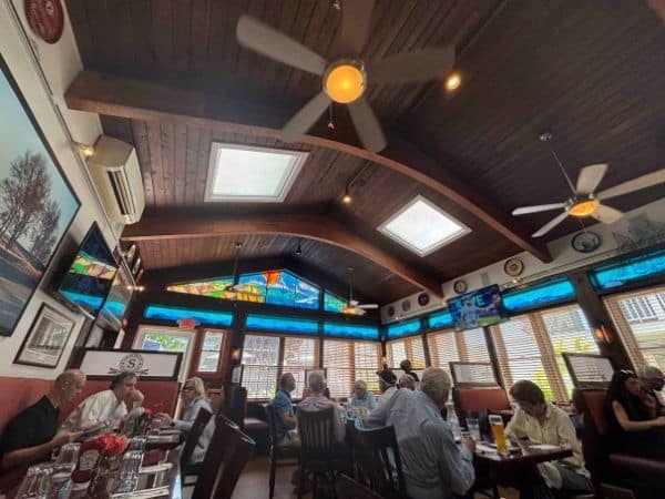 Entertainment options at the hamptons are endless. In this photo, inside the publick house is packed with people seated at their tables, surrounded on all sides by full length windows. The top of the windows aare stained glass, vibraating color into the high ceiling room, and a lrge ceiling fan hangs from the wooden ceiling. 